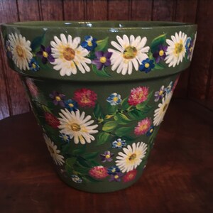 May include: A large, green ceramic flower pot decorated with hand-painted white daisies, purple, pink, and blue flowers. The floral design wraps around the pot, creating a vibrant and cheerful aesthetic. The pot is suitable for indoor or outdoor use.