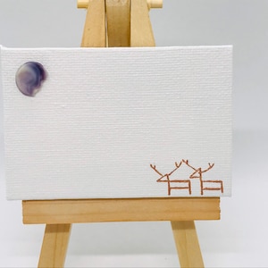 Könnte beinhalten: Ein kleines weißes Leinwandbild auf einer Staffelei mit einem lila und weißen Mond und zwei braunen Hirschsilhouetten.