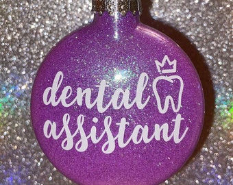 May include: A round, glittery purple Christmas ornament. The ornament has the words "dental assistant" in white script, along with a white crown and tooth graphic. The ornament has a silver metal cap and a black string for hanging.