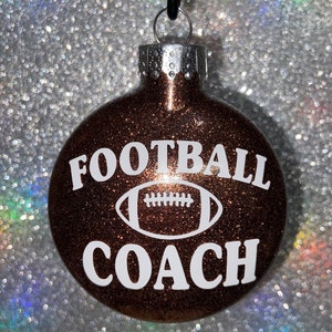 May include: A brown glitter Christmas ornament with the words "FOOTBALL COACH" in white, along with a white football graphic. The ornament has a silver metal cap and a black string for hanging.