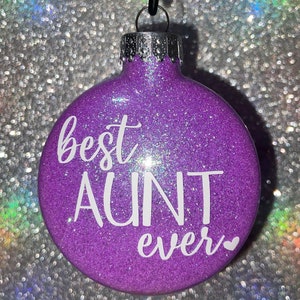 May include: A purple glitter Christmas ornament with the words "best AUNT ever" in white script. The ornament has a silver metal cap and a black string for hanging. A small white heart is at the end of the text.