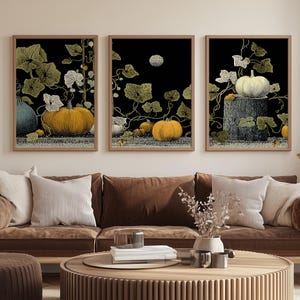Puede incluir: Tres impresiones artísticas enmarcadas con calabazas y follaje sobre un fondo negro. Las calabazas son de varios tonos de amarillo y naranja, con hojas y enredaderas verdes. La obra de arte se exhibe sobre un sofá marrón con cojines blancos.