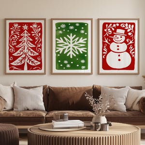 May include: Three framed Christmas-themed art prints. The prints feature a red Christmas tree, a green snowflake, and a red snowman, all with white designs. The artwork is displayed above a brown sofa in a living room setting.
