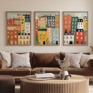 Könnte beinhalten: Drei gerahmte Kunstdrucke mit farbenfrohen Gebäuden mit roten, orangefarbenen, gelben und blauen Fassaden. Das Kunstwerk hängt über einem braunen Sofa mit weißen Kissen. Ein runder Couchtisch und ein Hocker aus Holz befinden sich vor dem Sofa.
