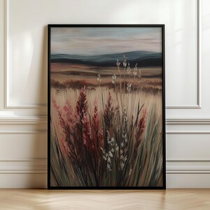 Op de afbeelding: Ingelijste kunstafdruk met een landschap met hoog gras en wilde bloemen in roze, beige en groene tinten. De achtergrond toont glooiende heuvels en een gedempte lucht. Het kunstwerk is ingelijst in een zwart frame.