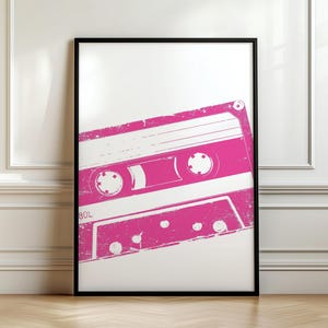 May include: A framed print featuring a distressed, pink cassette tape design. The cassette tape is set against a white background, with a black frame. The print is displayed on a wooden floor, leaning against a white wall.