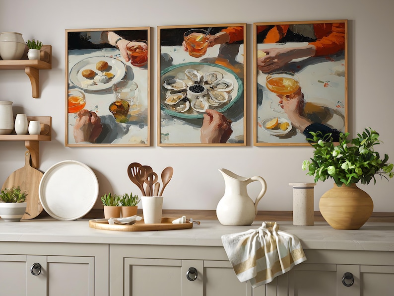 Pu&ograve; includere: Un trittico di stampe artistiche incorniciate raffiguranti una scena di ristorazione con mani che tengono bevande e piatti di cibo. Il piano della cucina espone una brocca bianca, un vassoio di legno con utensili e un vaso con verde.