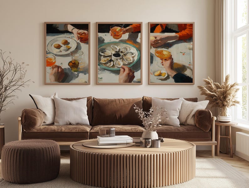 May include: Three framed art prints depicting a dining scene with hands, plates of food, and drinks. The artwork features a neutral color palette with brown, orange, and white tones. The prints are displayed above a brown sofa with white pillows.