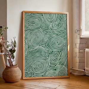 Puede incluir: Un cuadro enmarcado con un diseño abstracto verde y blanco de líneas onduladas. La obra de arte se muestra en un marco de madera natural y se apoya contra una pared blanca. El patrón crea una sensación de movimiento y profundidad.