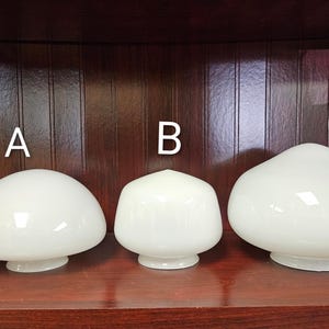 May include: Three white glass lamp shades of varying sizes are displayed on a dark wooden shelf. The shades have a rounded, mushroom-like shape, with a wider top and a narrower base. The shades are labeled A, B, and C.