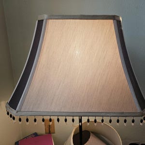 May include: A square-shaped lamp shade with a light beige fabric center and dark charcoal gray fabric panels on the corners. The bottom edge is decorated with a fringe of small, alternating black and clear beads hanging from the trim.