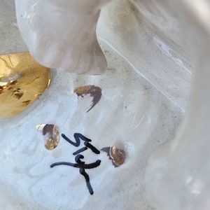 May include: White ceramic figurine with gold accents and a black signature. The figurine is of a woman with a flowing dress and a crown.