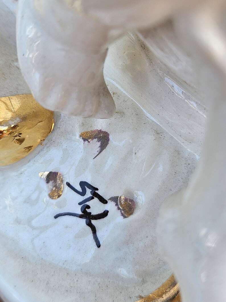 May include: White ceramic figurine with gold accents and a black signature. The figurine is of a woman with a flowing dress and a crown.
