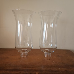 May include: Two clear glass hurricane lamp shades with a flared top and a cylindrical base. The shades are transparent, allowing light to pass through. They are placed on a wooden surface.