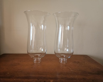 May include: Two clear glass hurricane lamp shades with a flared top and a cylindrical base. The shades are transparent, allowing light to pass through. They are placed on a wooden surface.
