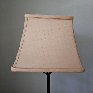 May include: A rectangular lampshade in a soft beige color with a subtle dotted pattern. The shade has a slightly flared shape and is trimmed with a darker beige border. The lamp base is black.