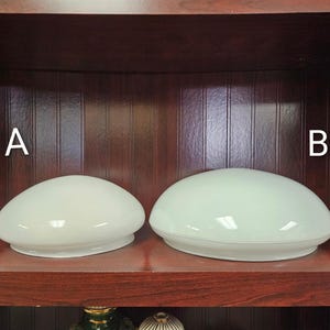May include: Two off-white, oval-shaped glass light fixtures, labeled A and B, sit on a dark wooden shelf. The fixtures have a smooth, glossy surface and a rounded, shallow design. The background is a dark wood cabinet.