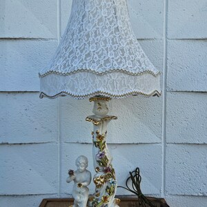 May include: A white porcelain table lamp with a cherub figurine and floral details. The lamp has a white lace lampshade.