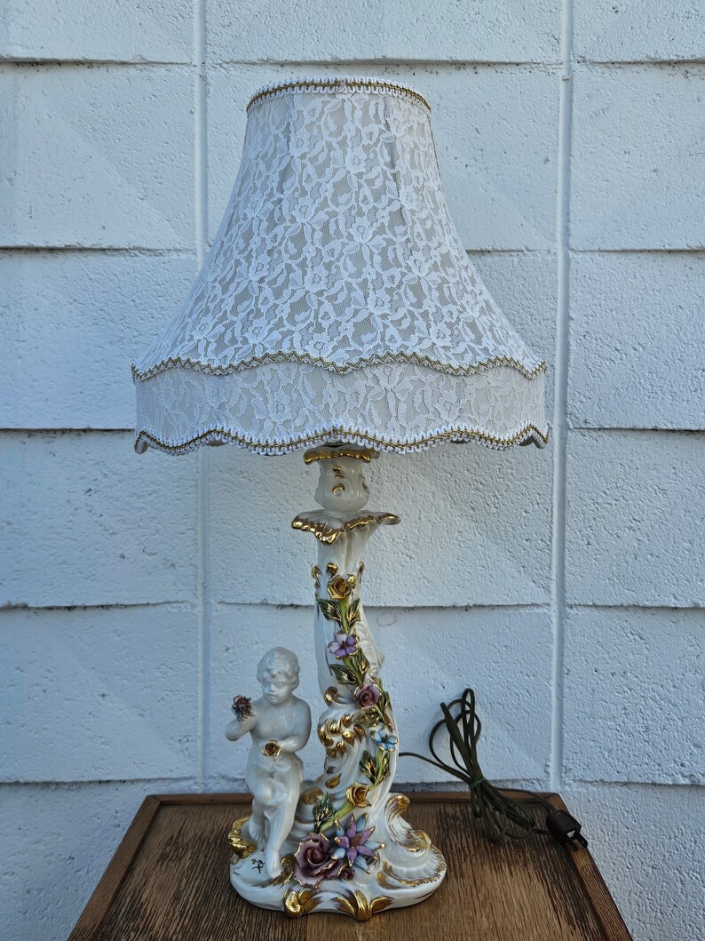 May include: A white porcelain table lamp with a cherub figurine and floral details. The lamp has a white lace lampshade.