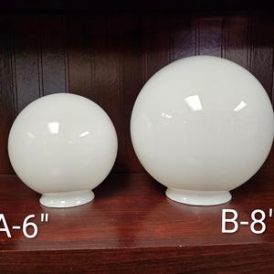 May include: Two white globe light fixtures, one labeled A-6" and the other B-8", sit on a dark wooden surface. The spherical shades have a glossy finish and sit on a small, circular base. The background is a dark wood.