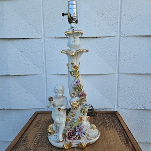 May include: A white porcelain table lamp with a cherub figurine and floral accents. The lamp base is decorated with gold accents and features a variety of flowers, including roses, lilies, and daisies.