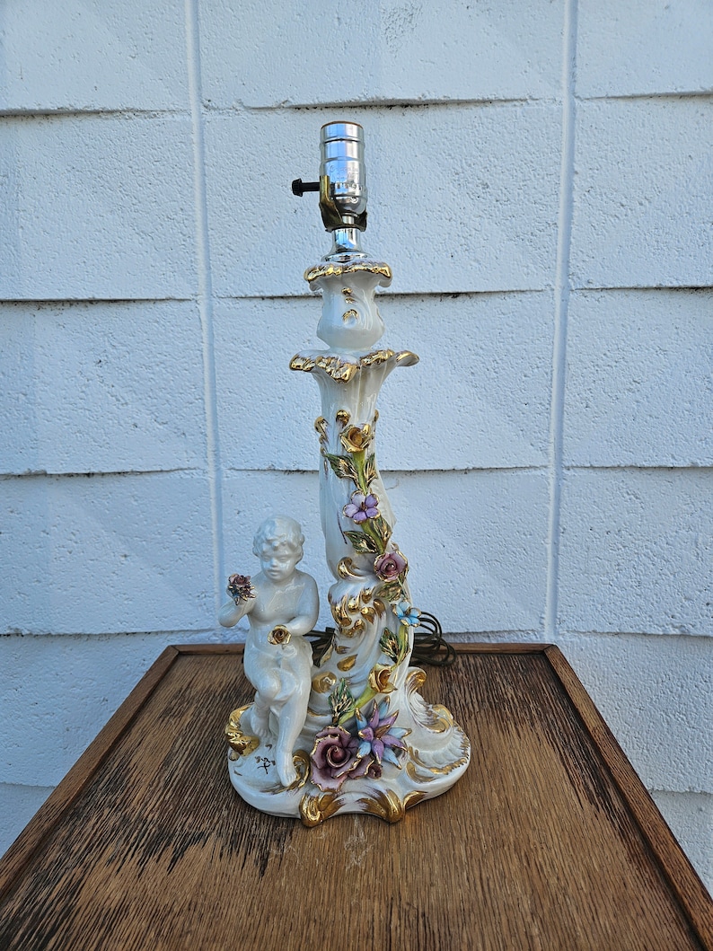 May include: A white porcelain table lamp with a cherub figurine and floral accents. The lamp base is decorated with gold accents and features a variety of flowers, including roses, lilies, and daisies.