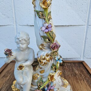 May include: A white porcelain lamp base with a cherub holding a rose. The base is decorated with gold accents and colorful flowers.