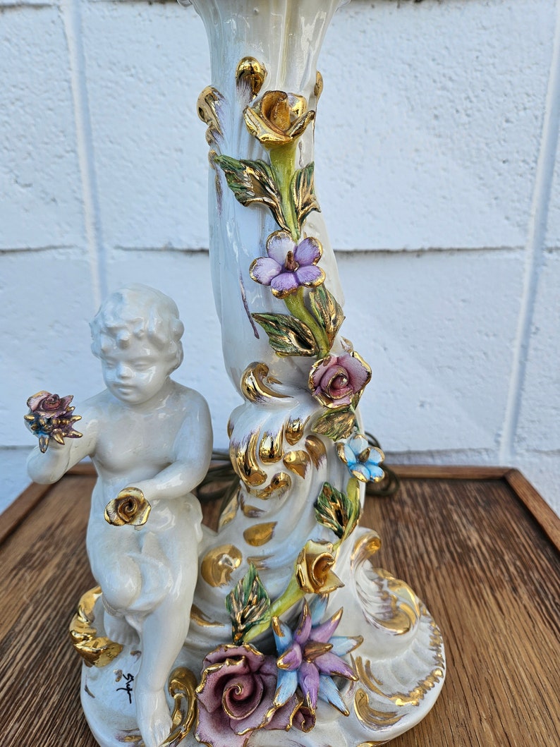 May include: A white porcelain lamp base with a cherub holding a rose. The base is decorated with gold accents and colorful flowers.