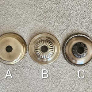 May include: Three antique brass ceiling canopy plates. Plate A is a simple, flat design. Plate B has a fluted pattern. Plate C features a rope-like edge and a central, ridged design. Each plate has a central hole.