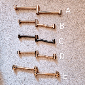 May include: Five different styles of gold and black metal towel bars. The bars are all about 12 inches long and have different shapes and curves. The bars are arranged vertically from top to bottom, with the letter A next to the top bar, B next to the second bar, and so on.