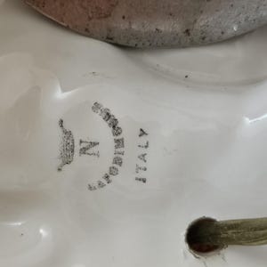 May include: Close-up of a white ceramic lamp base with a gold metal accent. The base has a black and white logo that reads "CAPODIMONTE ITALY" with a crown and the letter "N".
