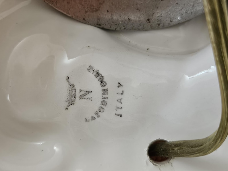 May include: Close-up of a white ceramic lamp base with a gold metal accent. The base has a black and white logo that reads "CAPODIMONTE ITALY" with a crown and the letter "N".