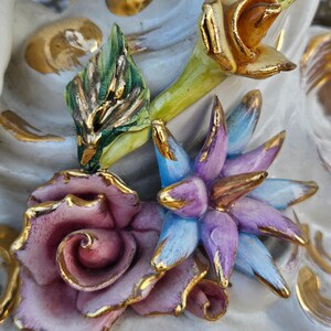 May include: Close-up of a porcelain figurine with detailed floral accents. The figurine features a pink rose, a blue flower, and a yellow flower with green leaves. The flowers are accented with gold paint.