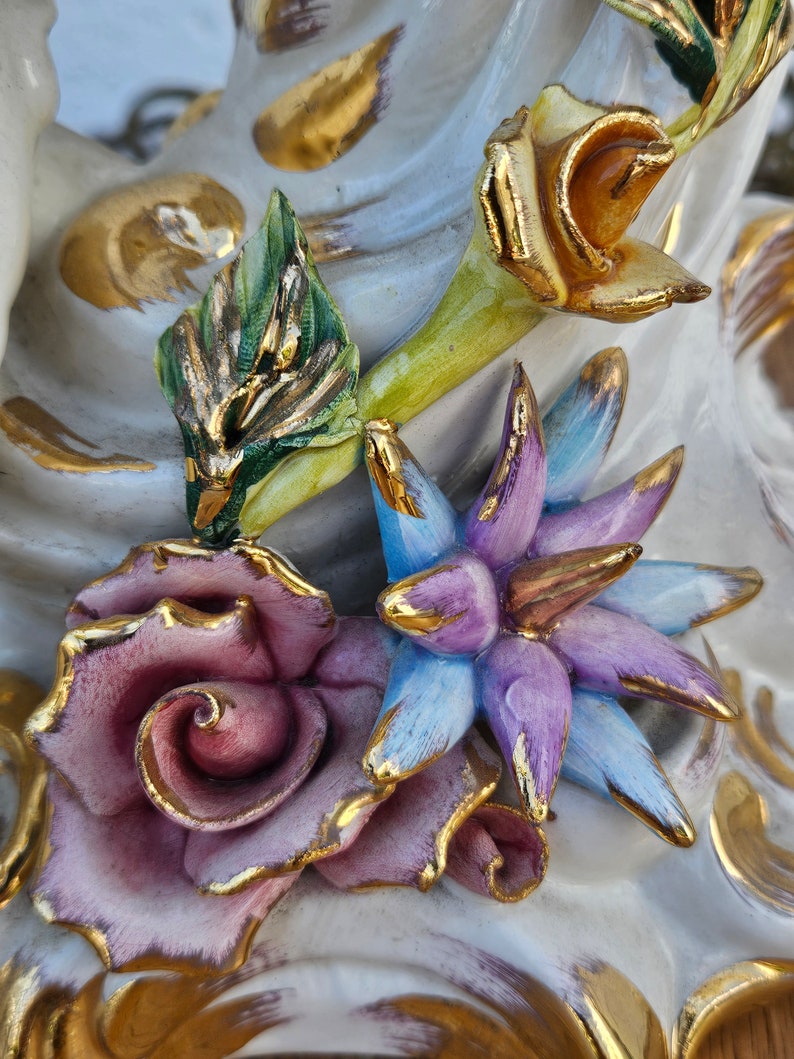 May include: Close-up of a porcelain figurine with detailed floral accents. The figurine features a pink rose, a blue flower, and a yellow flower with green leaves. The flowers are accented with gold paint.