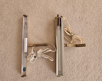 May include: Two vintage wall-mounted light fixtures. Each fixture has a rectangular, light-colored metal housing with a clear tube for the light bulb. The fixtures are connected by a brass-colored arm and have white cords.