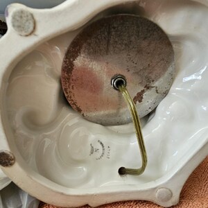 May include: Close-up of the bottom of a white ceramic lamp base with a metal lamp socket and a clear cord. The base has a decorative design and a label that reads "Made in Italy".