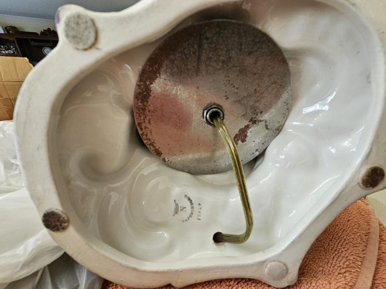 May include: Close-up of the bottom of a white ceramic lamp base with a metal lamp socket and a clear cord. The base has a decorative design and a label that reads "Made in Italy".