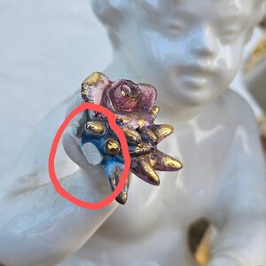 May include: A white porcelain figurine of a cherub holding a pink and gold rose with blue and gold accents. The cherub's face is painted with delicate features and the rose is detailed with realistic petals.