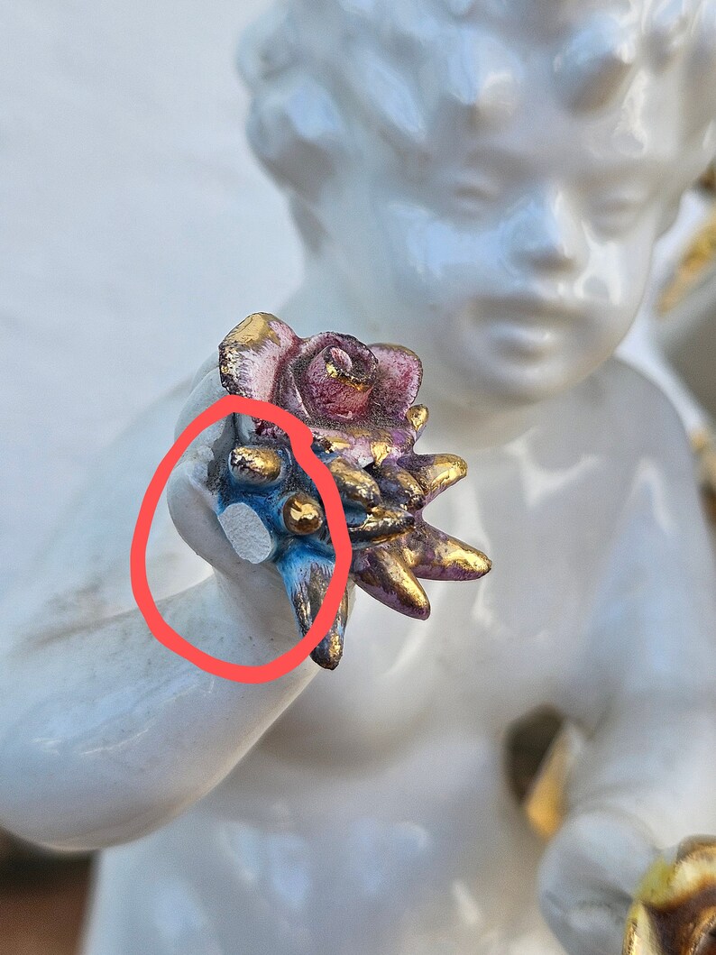 May include: A white porcelain figurine of a cherub holding a pink and gold rose with blue and gold accents. The cherub's face is painted with delicate features and the rose is detailed with realistic petals.
