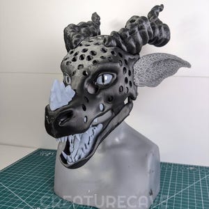 May include: A detailed, black and gray dragon mask with textured horns, ears, and a snout. The mask features open eye sockets, a row of sharp teeth, and a light blue accent on the nose. The mask is displayed on a gray mannequin head.