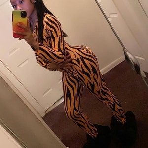 May include: A one-piece jumpsuit with a tiger stripe pattern in orange and black. The long-sleeved jumpsuit is paired with black fuzzy slippers. The person in the image is holding a colorful phone.