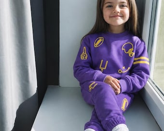 May include: A purple long-sleeve sweatshirt and matching sweatpants set featuring a yellow graphic print of American football icons including helmets, footballs, goal posts, and foam fingers, paired with white sneakers and white ribbed socks.
