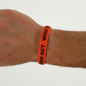 May include: Orange silicone wristband with the text "WHO DEY WHO" in black.
