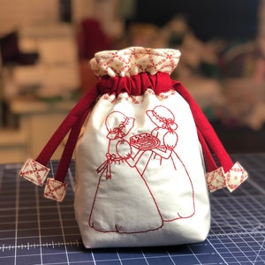 May include: White drawstring bag with red trim and embroidered red images of two girls holding a pie. The bag has red and white patterned fabric on the top and bottom.