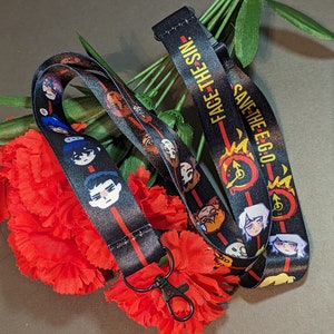Puede incluir: Cordón negro con caras de personajes de dibujos animados y el texto "FACE THE SIN." y "SAVE THE E.G.O." Una raya roja vertical recorre el centro. Se adjunta un cierre de metal negro. El cordón se muestra con flores rojas.