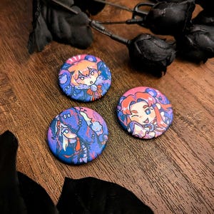 Puede incluir: Tres botones redondos y coloridos con ilustraciones de personajes de estilo anime. Los botones presentan tonos de azul, morado, rosa y naranja. Fondo de madera oscura con acentos de rosas negras.