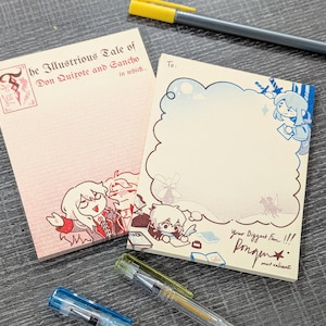 以下が含まれることがあります： イラストとテキストが入った2つのメモ帳。1つのメモ帳には「he Illustrious Tale of Don Quixote and Sancho in which」と書かれています。もう1つのメモ帳には「To: Your Biggest Fan!!! Pongn mut valiant」と書かれています。背景にはペンがあります。