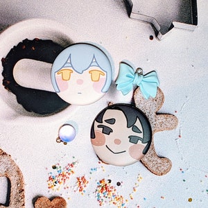 May include: Two round pins with cartoon faces. One pin is blue with a white face and the other is white with a brown face. The pins are on a white surface with a brown cookie and a blue bow.
