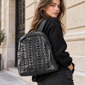 May include: A black leather backpack featuring a rounded top, a top handle, and adjustable shoulder straps. The front panel is decorated with vertical braided leather strips and rows of small, round, black studs arranged in a symmetrical pattern.