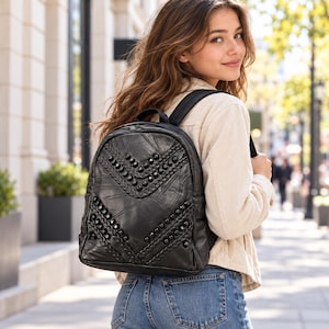 May include: A black leather backpack featuring a chevron pattern of braided leather strips and round black studs. The backpack has a rounded top, a top handle, and adjustable shoulder straps, with a visible zipper closure along the main compartment.